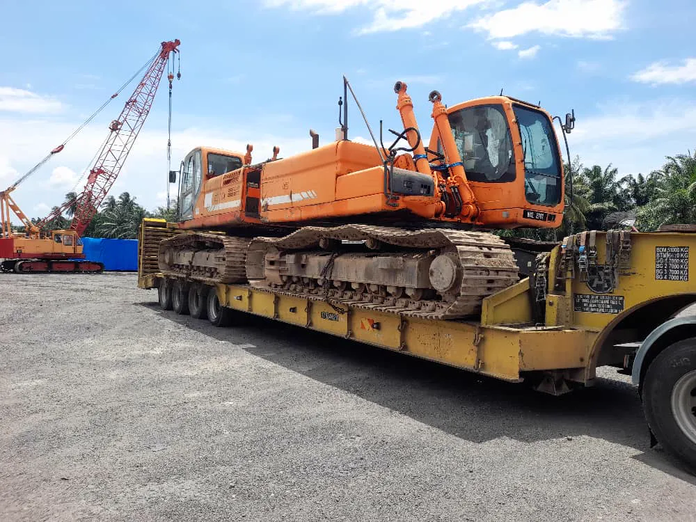 An orange excavator chained down on a yellow multi-axle lowloader, a crawler crane standing in the yard behind.