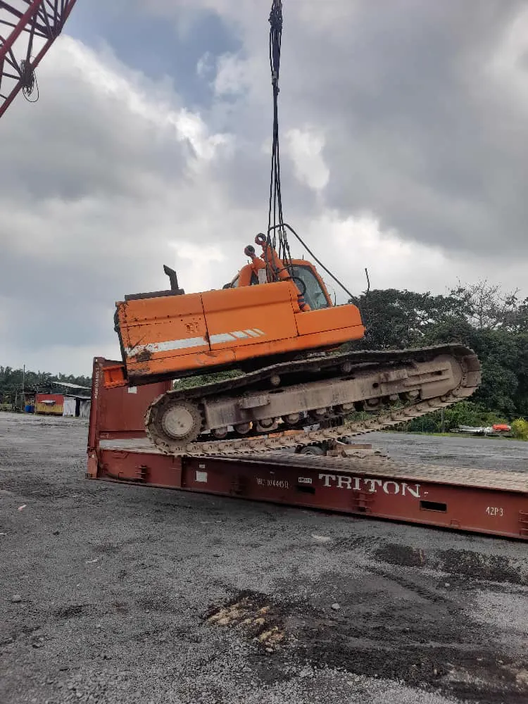 An excavator suspended from a crane hook, being lowered onto a flat rack container.