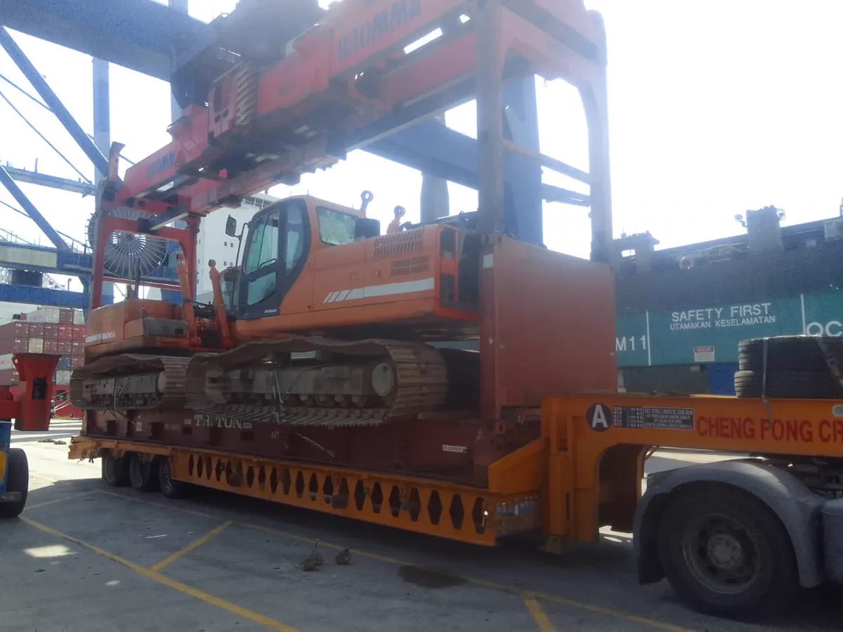 An excavator on a flat rack on a trailer, beneath container handling equipment in a port terminal.