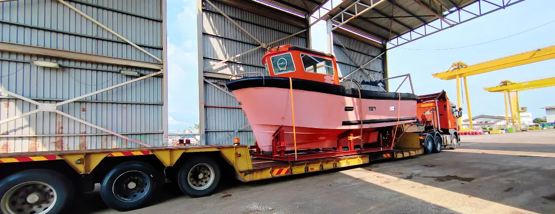 A boat hull secured on a lowloader trailer beside a covered yard, with yellow gantry cranes in the background.